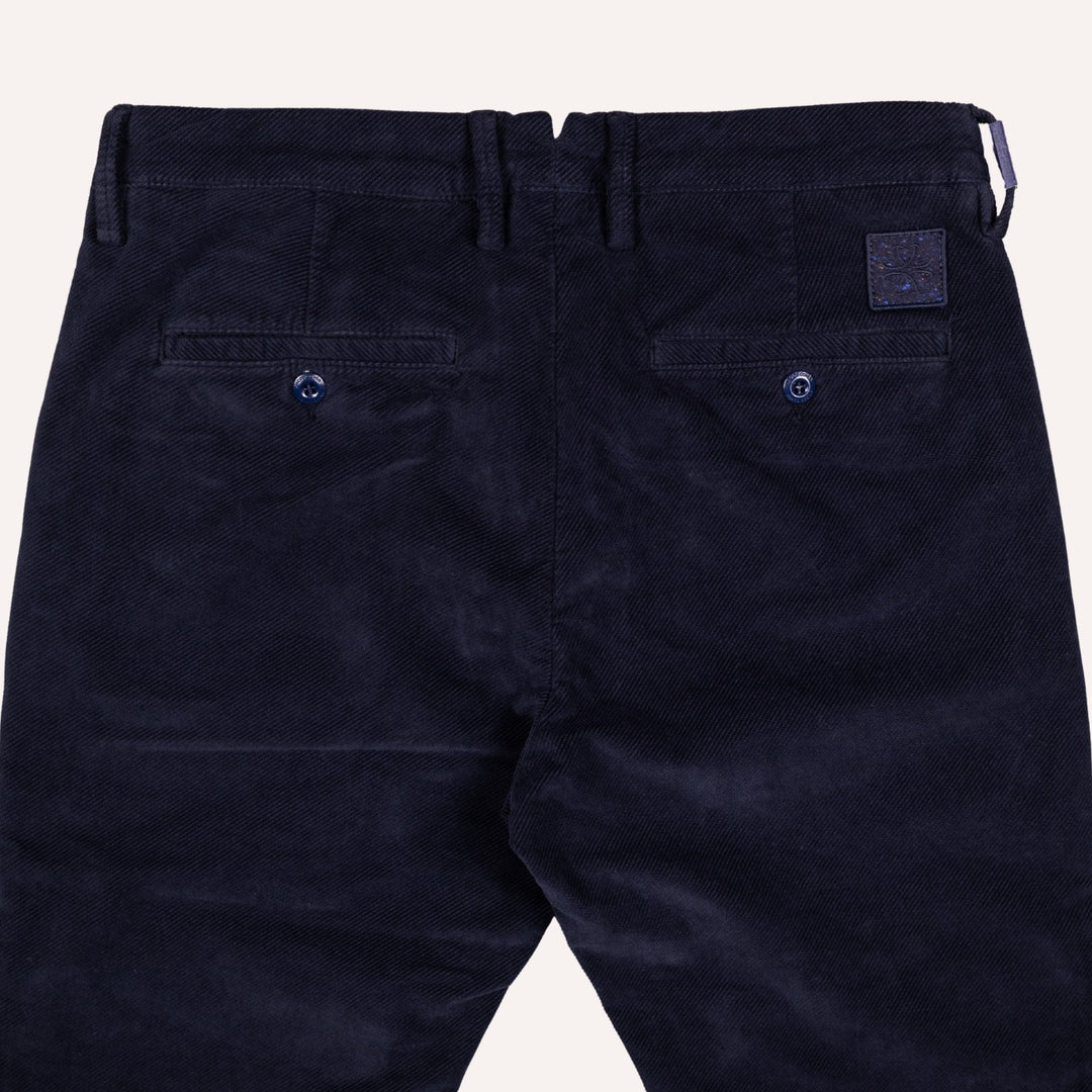 Close-up back view of dark navy twill trousers showing the waistband, belt loops, two buttoned back pockets, and a small square brand patch on the upper right side.