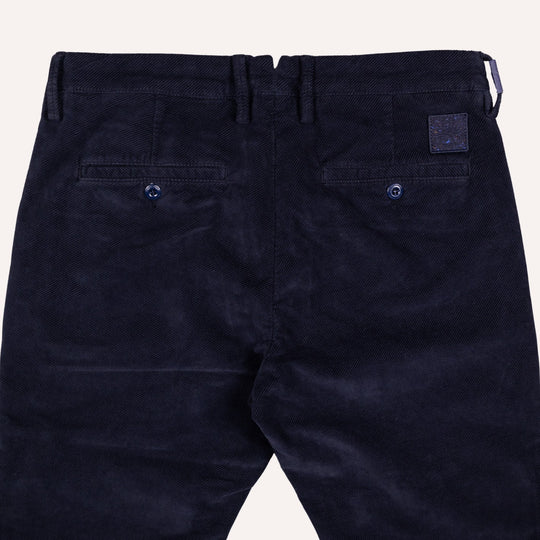 Close-up back view of dark navy twill trousers showing the waistband, belt loops, two buttoned back pockets, and a small square brand patch on the upper right side.