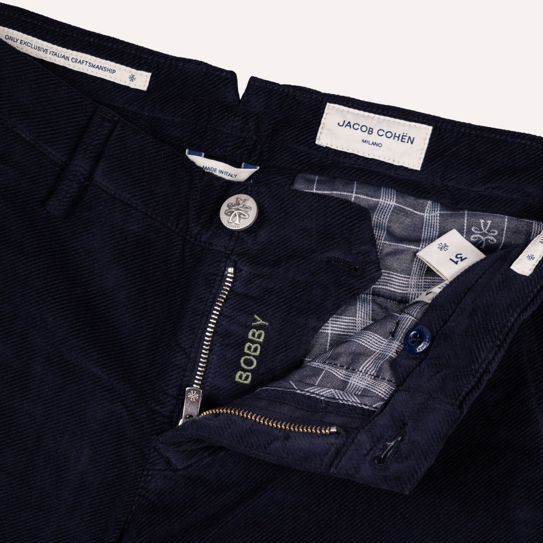 Close-up of dark navy twill trousers by Jacob Cohen, showing the waistband, button closure, zippered fly with the brand name BOBBY embroidered in gold, and a plaid inner lining.