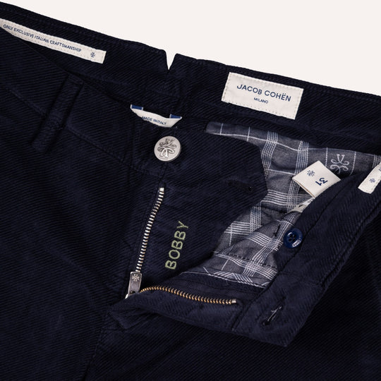 Close-up of dark navy twill trousers by Jacob Cohen, showing the waistband, button closure, zippered fly with the brand name BOBBY embroidered in gold, and a plaid inner lining.