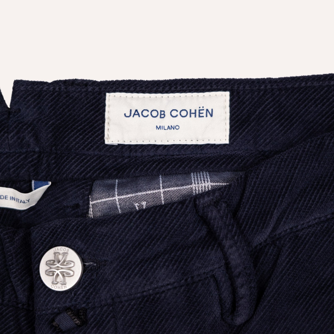 Close-up of dark navy blue twill pants showing the interior waistband, a white fabric label reading JACOB COHÉN MILANO, and a silver-tone button with a logo.