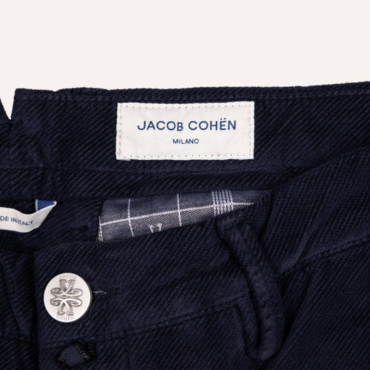 Close-up of dark navy blue twill pants showing the interior waistband, a white fabric label reading JACOB COHÉN MILANO, and a silver-tone button with a logo.