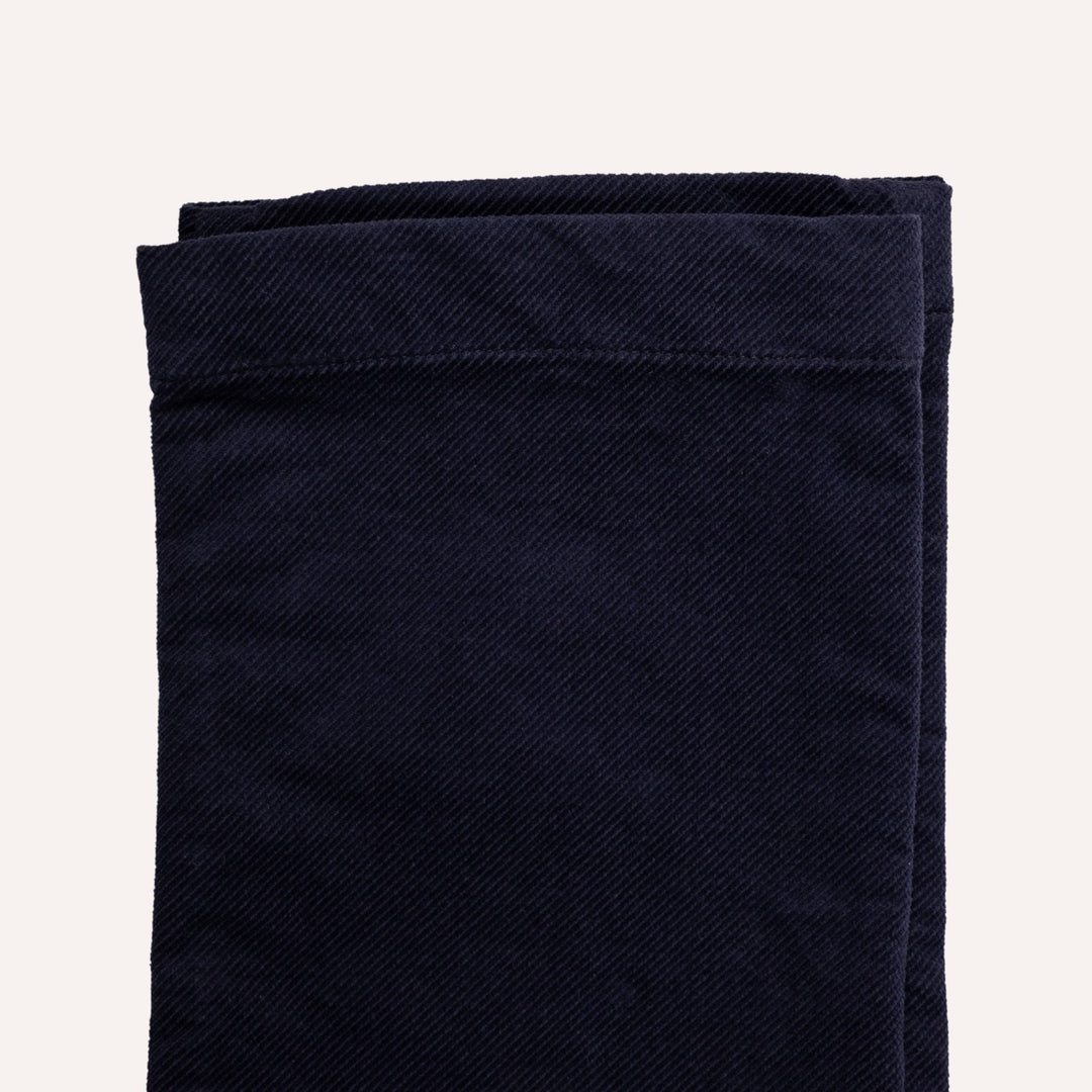 A close-up of folded dark navy blue fabric, showing a textured twill weave and neat hem stitching, set against a solid off-white background.