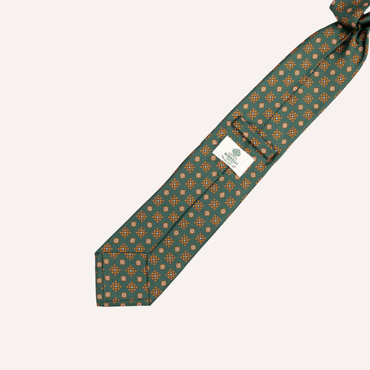 A dark green silk necktie with a repeating orange and yellow geometric pattern, laid diagonally on a light beige background. A white brand label is visible on the main body of the tie.