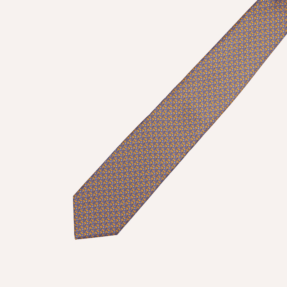 A patterned necktie with a repeating geometric design in brown and blue on a white base, shown diagonally on a light cream background.