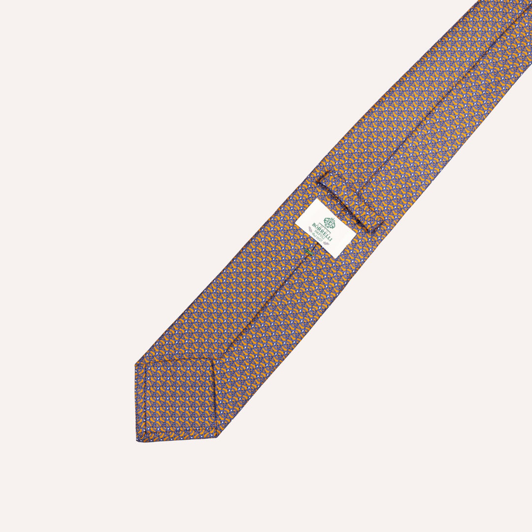 A close-up of a patterned necktie laid diagonally on a light cream background. The tie features a repeating geometric design in orange and blue on a dark base, with a small white brand label sewn near the tip.