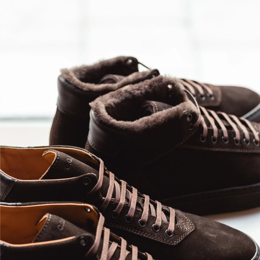 A pair of dark brown suede high-top lace-up boots with plush brown faux-fur lining, shown in a close-up studio-style shot against a bright white background.
