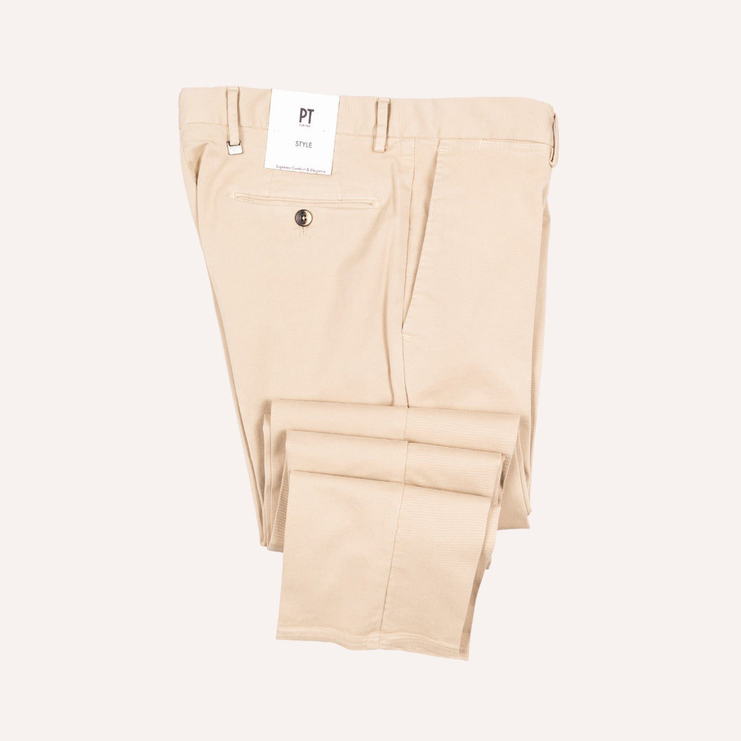 A pair of light beige chino pants folded neatly, showing the back waistband, belt loops, a back pocket with a button, and a white brand tag that reads PT. The background is a solid, very light off-white color.