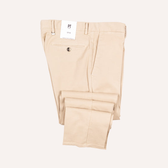 A pair of light beige chino pants folded neatly, showing the back waistband, belt loops, a back pocket with a button, and a white brand tag that reads PT. The background is a solid, very light off-white color.