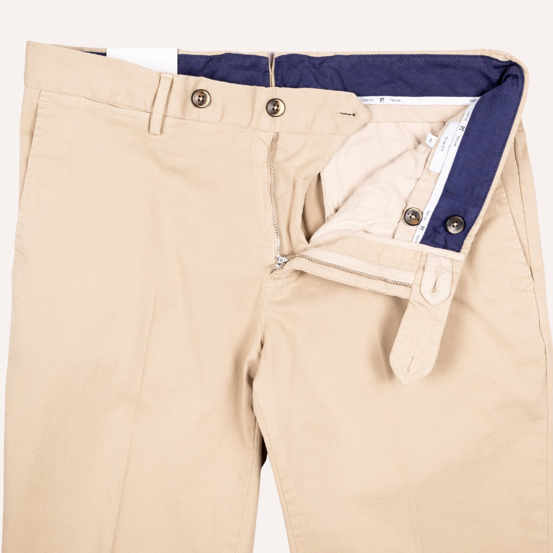 Beige chino-style pants shown folded flat with the front opening and interior waistband visible, including a navy inner lining and metal buttons.