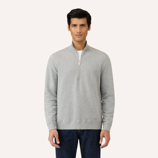 A man modeling a light gray quarter-zip pullover sweater with a stand collar, worn over a white undershirt and paired with dark blue jeans, against a solid off-white background.