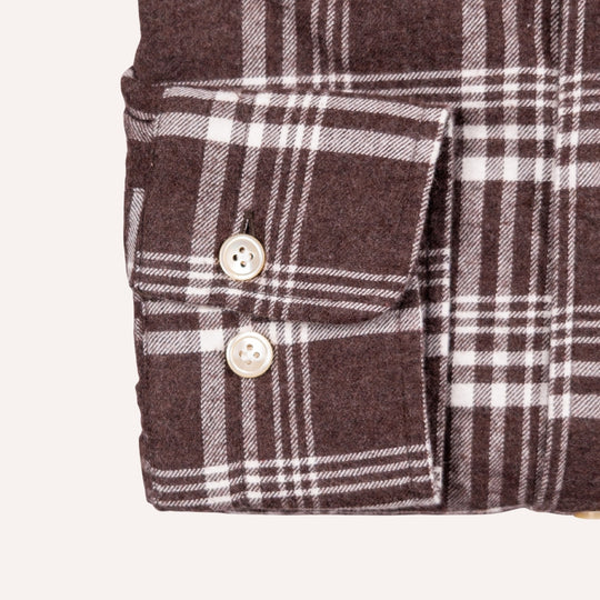 Close-up of a brown and white plaid garment showing a buttoned pocket flap and fabric texture on a light neutral background.
