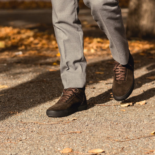 A person wearing brown suede lace-up shoes and gray trousers walking outdoors on a gravel path with fallen leaves.